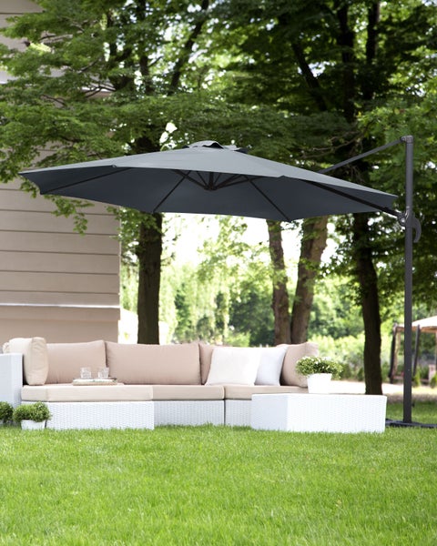 Großer grauer Ampelschirm über einer weißen Garten-Lounge-Garnitur aus Geflecht auf einer grünen Rasenfläche vor einem Waldhintergrund.