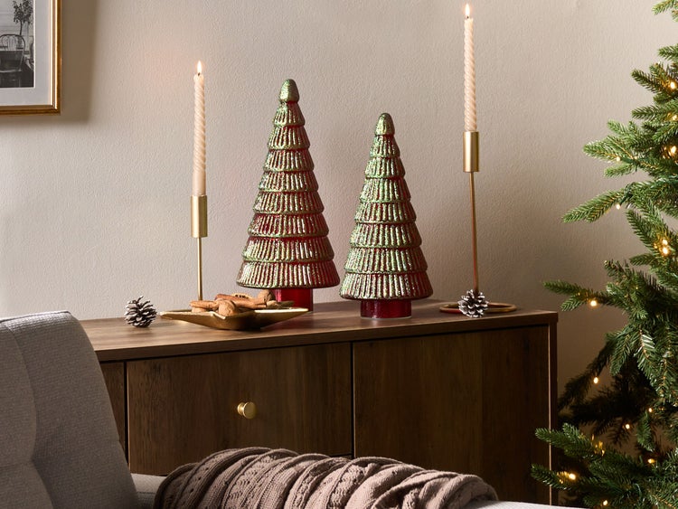 Festliche Szene mit Dekoration: Zwei Weihnachtsbäume, Kerzen, Tannenzapfen und eine Schale mit Zimtstangen auf einem Holzschrank neben einem Weihnachtsbaum.