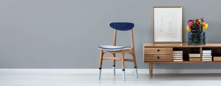 Hell gestalteter Wohnbereich mit Holzstuhl mit blauer Sitzfläche, Sideboard aus Holz, Bilderrahmen und Blumen in einer Vase.