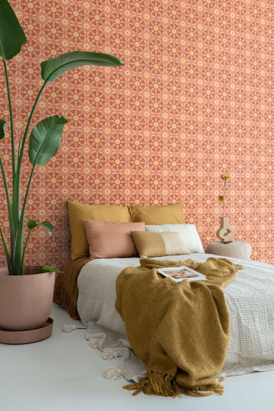 Hell gestaltetes Schlafzimmer mit gemusterter Vliestapete im Retro Design, Bett mit Kissen und Decke sowie einer großen Zimmerpflanze im Topf.