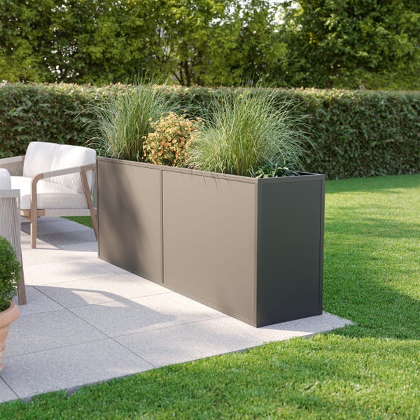 Rechteckiger Metall-Pflanzkübel in Anthrazit auf einer Terrasse, bepflanzt mit Gräsern, neben einer Rasenfläche und Gartenmöbeln.