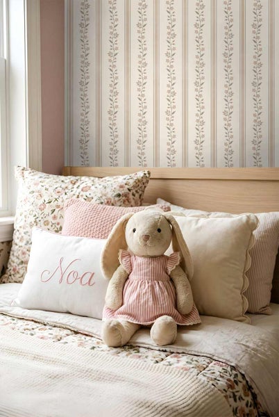 Tapete im Kinderzimmer mit Streifenmuster und floralen Girlanden, kombiniert mit passenden Kissen und einem Stofftier auf einem Holzbett.
