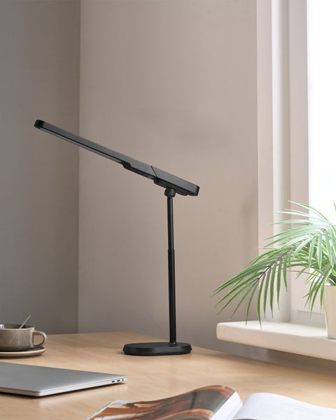 Schreibtischszene mit einer schwarzen Schreibtischlampe, Laptop, Tasse und einer Zimmerpflanze auf einer Holzoberfläche.