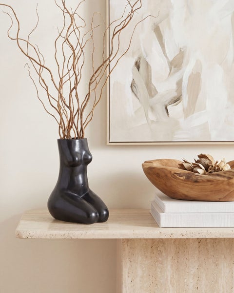 Moderne Dekoration mit schwarzer Vase in Torsoform, getrockneten Zweigen, Holzschale auf Büchern und abstraktem Bild auf einem Steintisch.