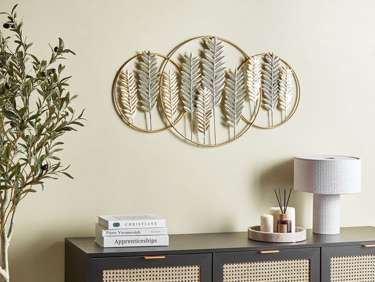 Dekoratives Wandobjekt mit goldenen Kreisen und stilisierten Blättern über einem Schrank mit Lampe und Dekoartikeln