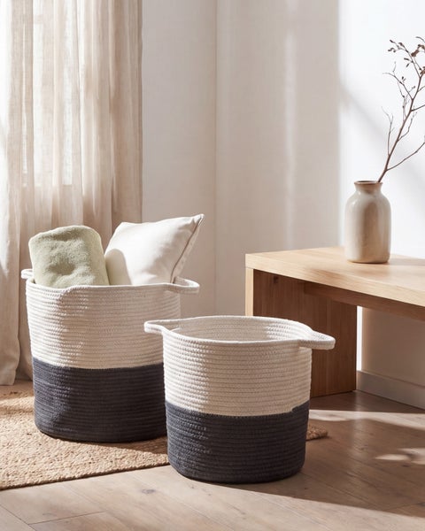 Zwei geflochtene Aufbewahrungskörbe aus Baumwolle in einem hellen Zimmer mit Holzbank und Jute-Teppich. Ein Korb ist mit Kissen und Decke gefüllt.