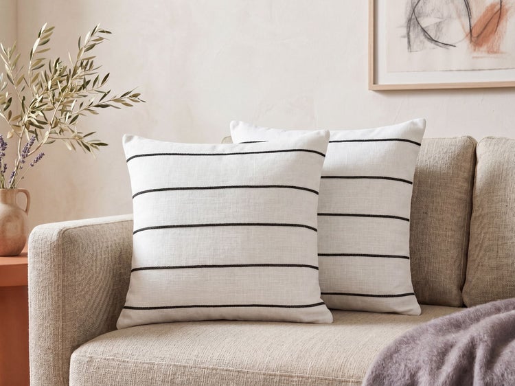 Zwei weiße Kissen mit schwarzen horizontalen Streifen in Leinenoptik auf einem hellen Sofa in einem hellen Wohnzimmer.