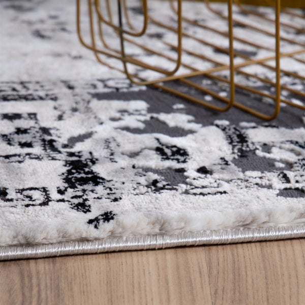 Nahaufnahme eines Teppichs mit weiß-grau-schwarzem Vintage-Muster, weicher Textur und geketteltem Rand auf Holzboden.