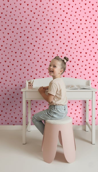 Mädchen sitzt an einem Schreibtisch vor einer Wand mit Herzmuster.