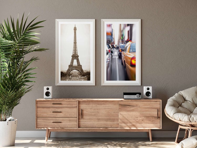 Modernes helles Wohnzimmer mit Sideboard aus Holz, zwei gerahmten Bildern vom Eiffelturm und einer Stadtszene, Zimmerpflanze und Korbsessel.