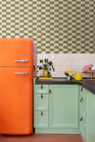 Küche mit mintgrünen Schränken, Ledergriffen, schwarzer Arbeitsplatte, weißen Fliesen, gemusterter Tapete und orangefarbenem Kühlschrank mit Smeg Logo.