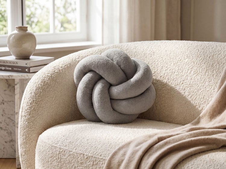 Graues Dekokissen in Knotenform aus weichem Plüsch auf einem hellen Bouclé-Sessel in einem hellen Wohnzimmer.