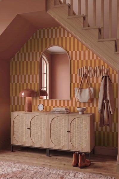 Heller Flur unter einer Treppe mit Sideboard aus Holz, Rattantüren, Rundbogenspiegel und geometrischer Tapete.