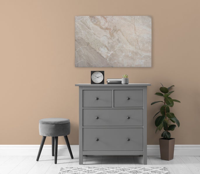 Graue Kommode mit drei Schubladen neben einem Hocker und einer Pflanze vor einer Wand mit einem abstrakten Wandbild.