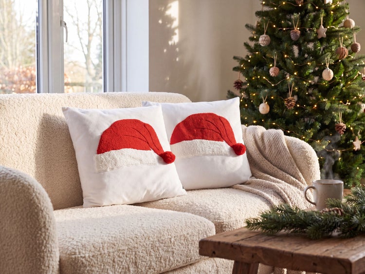 Zwei weiße Kissen mit Weihnachtsmützen-Motiv auf einem beigen Fleece-Sofa neben einem geschmückten Weihnachtsbaum in einem hellen Wohnzimmer.