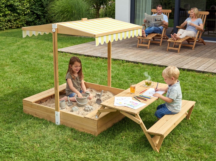 Holzsandkasten mit gelb-weiß gestreiftem Dach und integriertem Picknicktisch im hellen Garten, Kinder spielen im Sand und malen am Tisch.