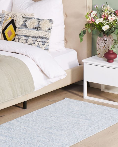 Modernes Schlafzimmer mit hellblauem Teppich, gepolstertem Bett, weißem Nachttisch und Blumendekoration auf Holzfußboden.