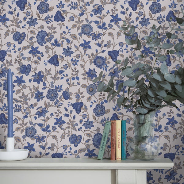 Vliestapete mit floralem Muster aus blauen Blumen, davor ein Kaminsims mit Kerze, Büchern und einer Vase mit Eukalyptus.