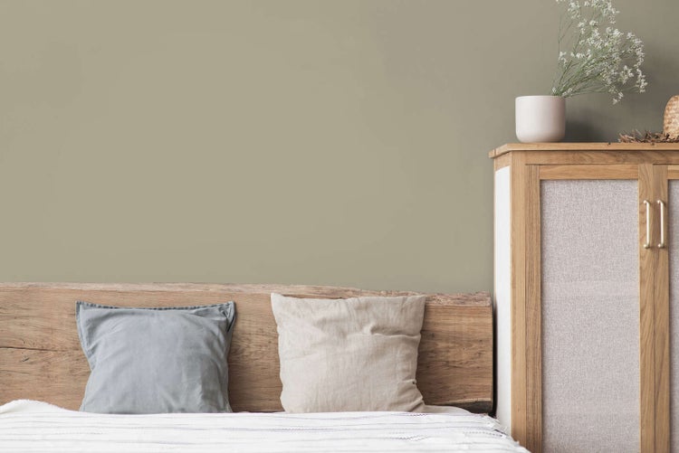 Schlafzimmerszene mit matt gestrichener Wand, einem Echtholz-Kopfteil mit Kissen und einem Holzschrank mit Textileinsätzen.