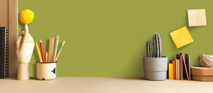 Organisierter Arbeitsplatz mit Schreibtisch, hölzerner Gliederhand, Pinseln, Stiften, Kaktus im Topf, Büchern und Notizzetteln an der Wand.