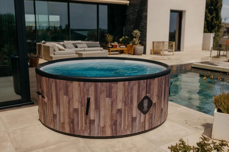 Runder aufblasbarer Whirlpool in Holzoptik mit Coco Spa Logo auf einer Terrasse neben einem Swimmingpool und Gartenmöbeln.