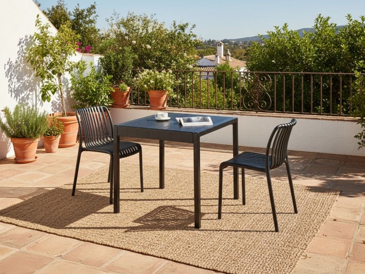 Schwarze Gartenmöbelgruppe mit quadratischem Tisch und zwei Stühlen auf einem Webteppich auf einer sonnigen Terrasse mit Pflanzen.