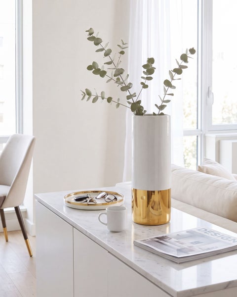Eine hohe zylindrische Vase mit einer Kombination aus weißer Oberfläche und metallisch glänzendem Sockel, gefüllt mit Eukalyptuszweigen.
