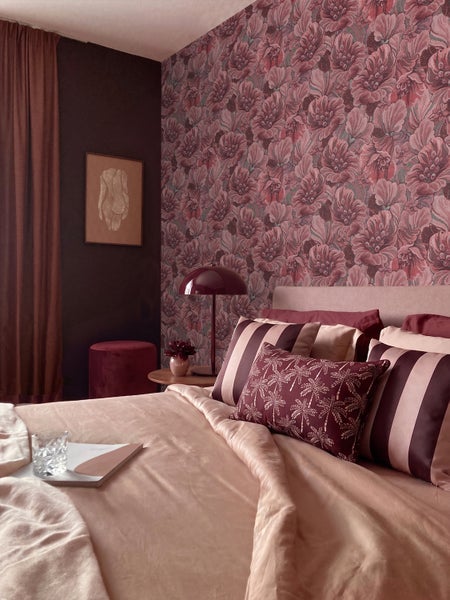 Schlafzimmer mit floral gemusterter Tapete, einem Bett mit Kissen in Streifen und Palmenoptik sowie einer pilzförmigen Tischleuchte.