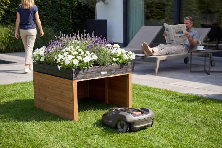 Moderner Garten mit Mähroboter-Garage aus Holz mit integriertem Pflanzkasten, ein Mähroboter auf dem Rasen und Personen auf der Terrasse.