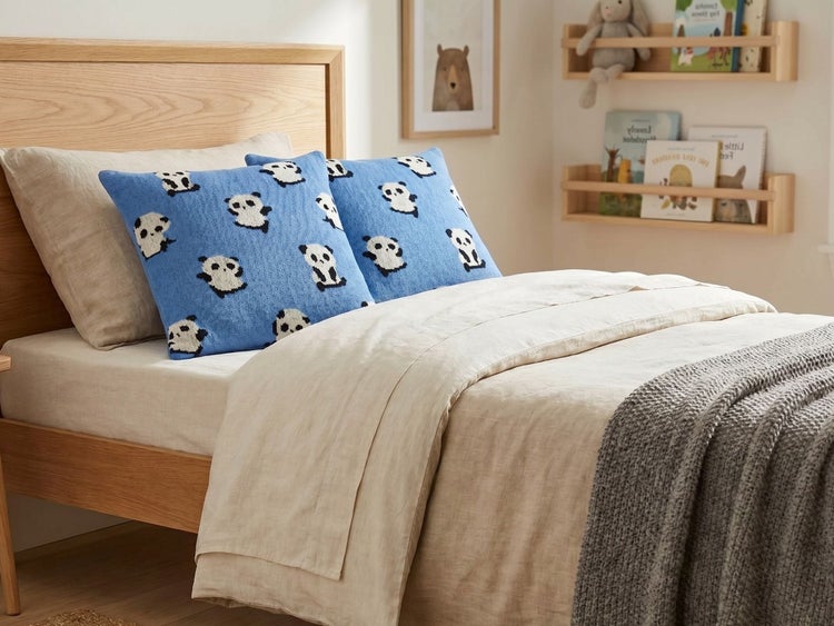 Kinderbett aus Holz mit hellem Bettzeug und blauen Zierkissen mit Panda-Motiv. Im Hintergrund hängen Holzregale mit Kinderbüchern an der Wand.