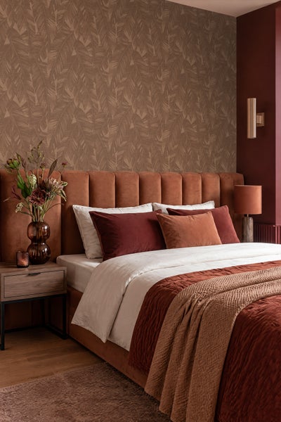 Modernes Schlafzimmer mit gepolstertem Bett, gemusterter Tapete mit Blattmotiven, dunklen Wohntextilien, Holz-Nachttisch und Wandleuchte.