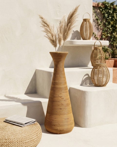 Hohe konische Bodenvase aus Rattangeflecht mit Pampasgras sowie passende Windlichter auf hellen Steinstufen.