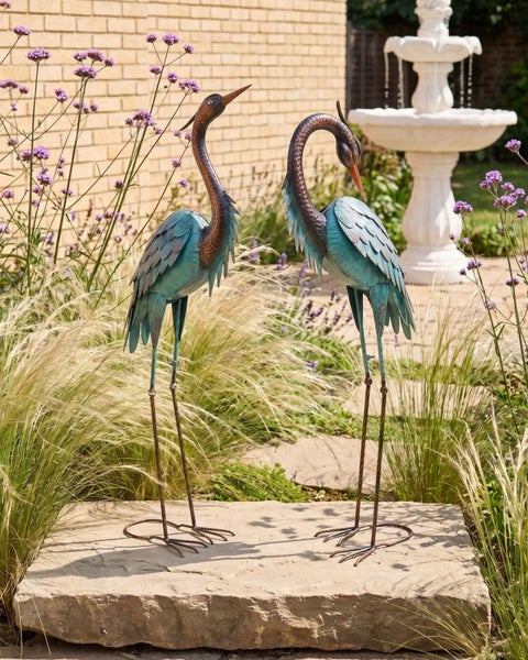 Zwei dekorative Reiher aus Metall auf einer Steinplatte in einem hellen Garten mit Ziergräsern, Blumen und einem Springbrunnen.
