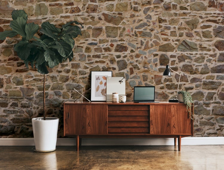 Fototapete in Natursteinoptik in einem Wohnzimmer mit hölzernem Sideboard, großer Zimmerpflanze und Schreibtischlampe.