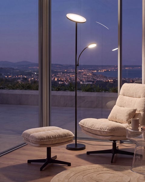 Moderne LED Deckenfluter Stehleuchte mit Lesearm neben einem Sessel mit Hocker vor einer großen Fensterfront mit nächtlichem Stadtblick.