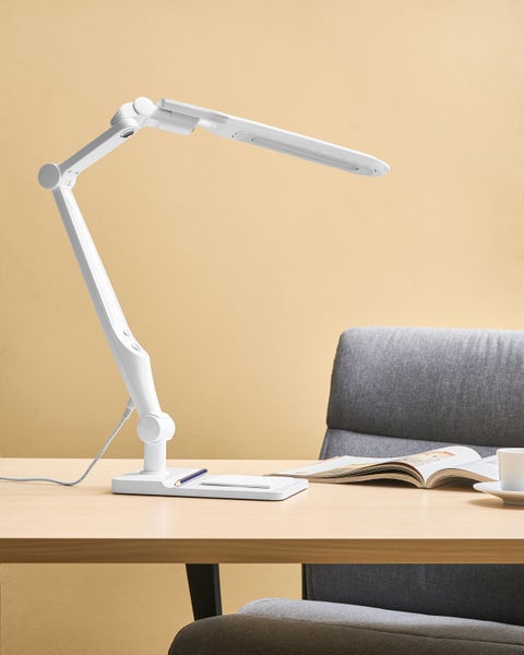 Verstellbare, weisse Schreibtischlampe auf einem Schreibtisch mit Stuhl, Buch, Tasse und Bleistift.