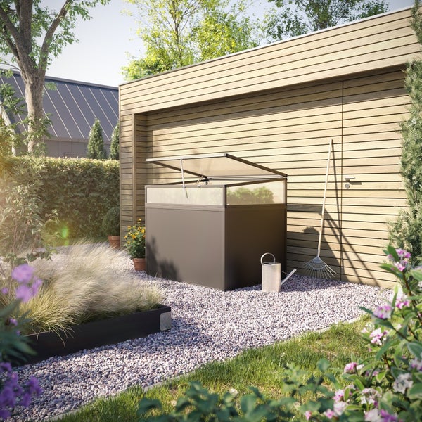 Frühbeet aus Metall und Polycarbonat in einem hellen, organisierten Garten vor einer Holzwand mit Gießkanne und Rechen.