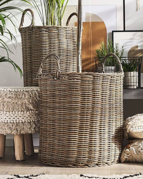 Zwei große, runde Flechtkörbe aus Rattan mit Griffen, arrangiert neben einem Holzhocker mit Fransenoberseite und verschiedenen Zimmerpflanzen.