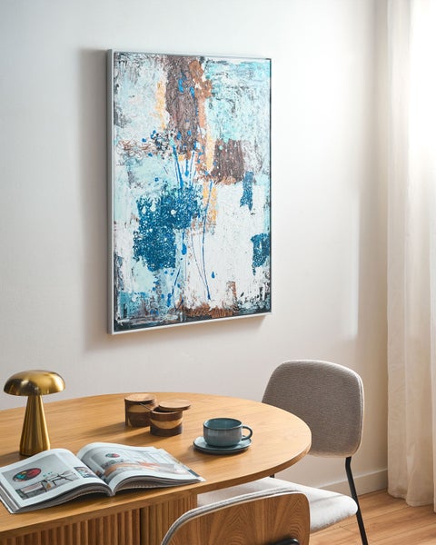 Modernes Wandbild mit abstraktem Motiv im silbernen Rahmen über einem runden Holztisch mit goldener Tischleuchte und Stuhl.