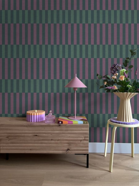 Vliestapete mit geometrischem Blockstreifenmuster in Grün und Lila hinter einer Holzkommode und einem Blumenständer.