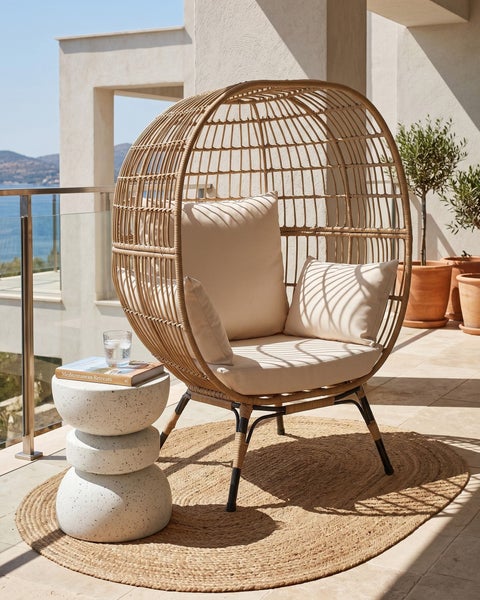 Moderner Gartenstuhl in Eiform aus hellem Geflecht mit hellen Polstern auf einer Terrasse. Daneben ein weißer Beistelltisch auf einem Jute-Teppich.