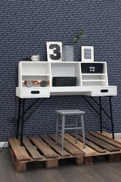 Szene mit einem weißen Schreibtisch mit Schubladen, Ablageflächen und Dekoration sowie einem grauen Hocker auf einer Holzpalette vor einer gemusterten Tapete.