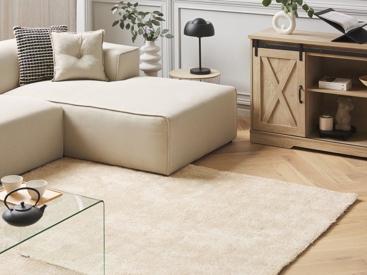 Modernes Wohnzimmer mit hellem Ecksofa, Teppich, Glastisch und Sideboard im Landhausstil mit Schiebetür vor einer Wand mit Zierleisten.