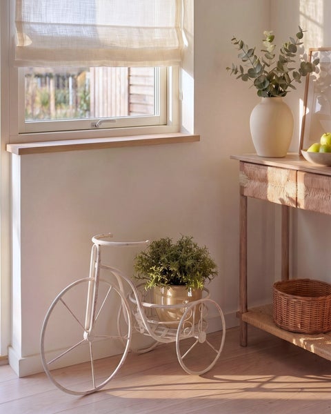Hell gestaltete Zimmerecke mit Fenster, Raffrollo, einem metallenen Blumenständer in Fahrradform und einem Konsolentisch aus Holz mit Korb.