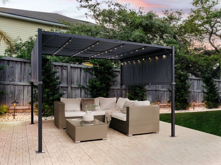 Moderne Pergola aus Metall mit LED-Beleuchtung über einer Lounge-Ecke aus Polyrattan auf einer gepflasterten Terrasse im Garten.