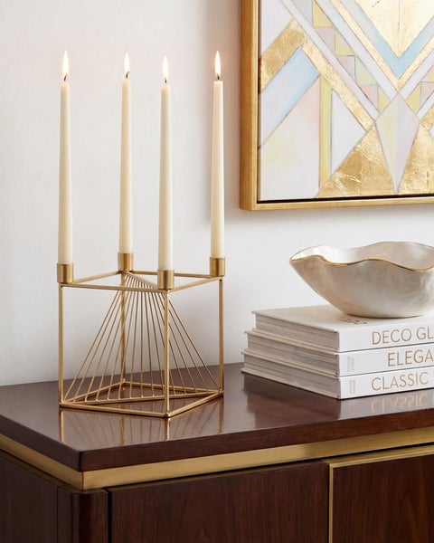 Geometrischer Kerzenständer aus Metall mit vier brennenden Kerzen auf einem Sideboard aus Holz neben Büchern, einer Schale und einem Wandbild.