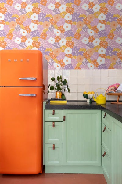 Eine bunte Küche im Retro-Stil mit geblümter Tapete, einem orangefarbenen Kühlschrank und grünen Schränken mit Ledergriffen.