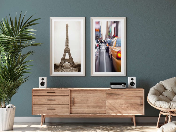 Modernes Wohnzimmer mit einem Sideboard aus Holz, zwei großen eingerahmten Wandbildern von Paris und New York sowie einer großen Zimmerpflanze.