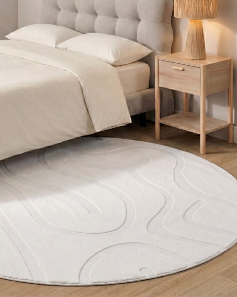 Runder Teppich mit abstraktem Reliefmuster vor einem gemütlichen Schlafzimmerbett mit Nachttisch und Tischlampe auf einem Holzboden.
