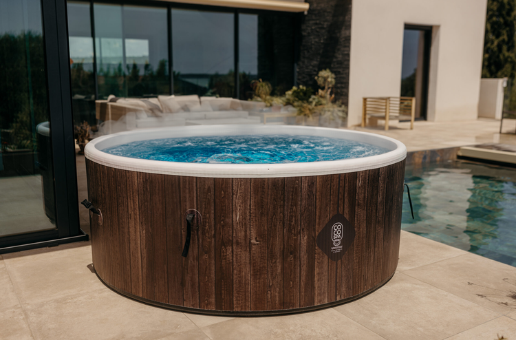 Runder Whirlpool mit Verkleidung in Holzoptik und COCO SPA Logo auf einer hellen Terrasse neben einem Pool.